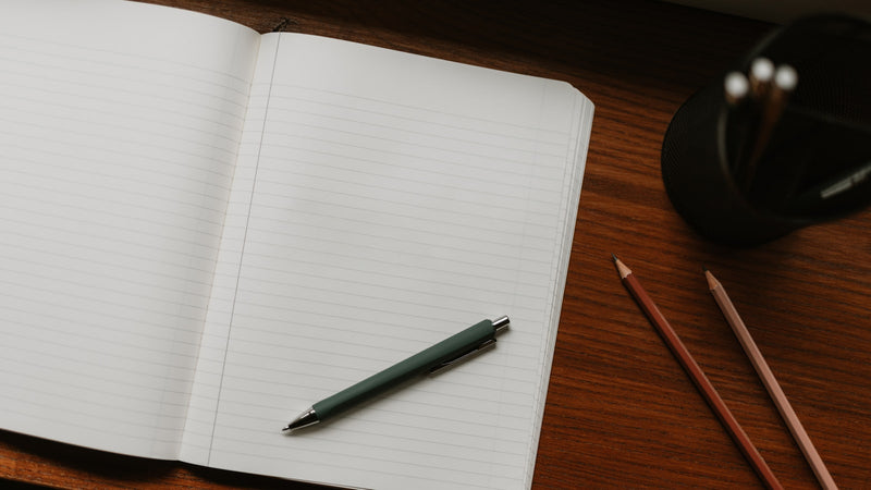 Open notebook with a pen and pencils on a wooden desk. Photo by Clay Banks on Unsplash.
